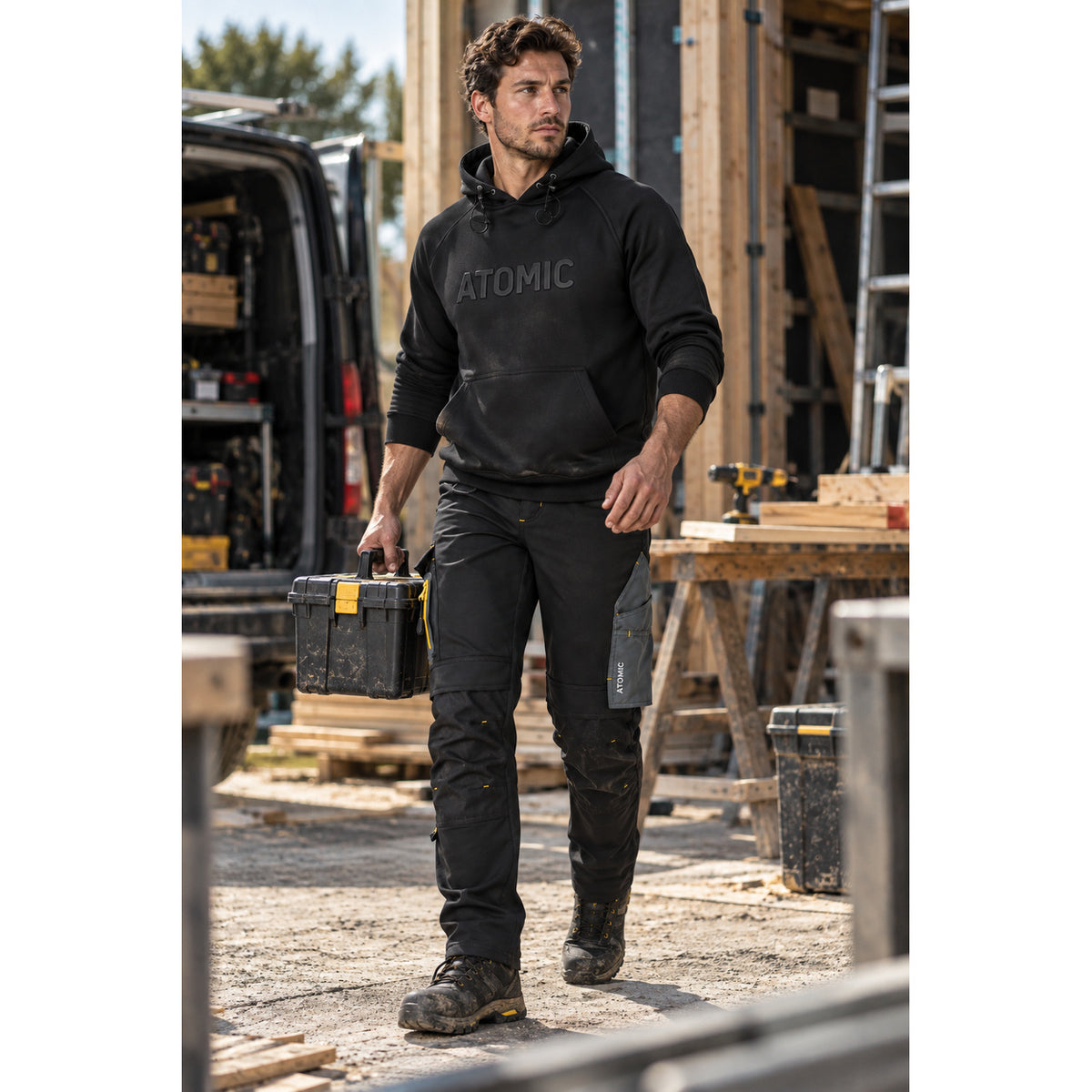 Tradesman wearing black ATOMIC work trousers and logo hoodie while carrying a toolbox on a construction site.