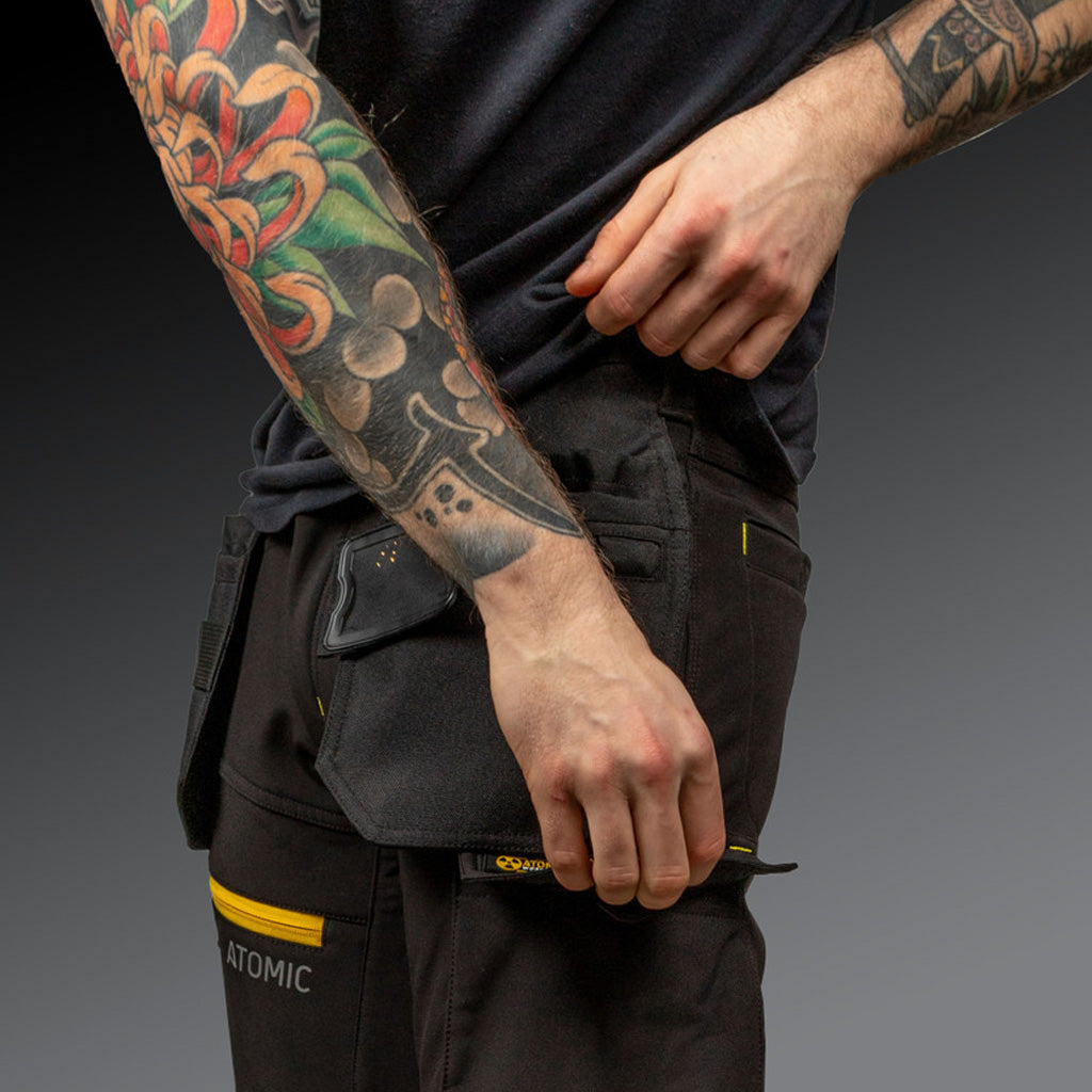 Person wearing black pants with a visible brand logo, holding a black jacket with pockets.