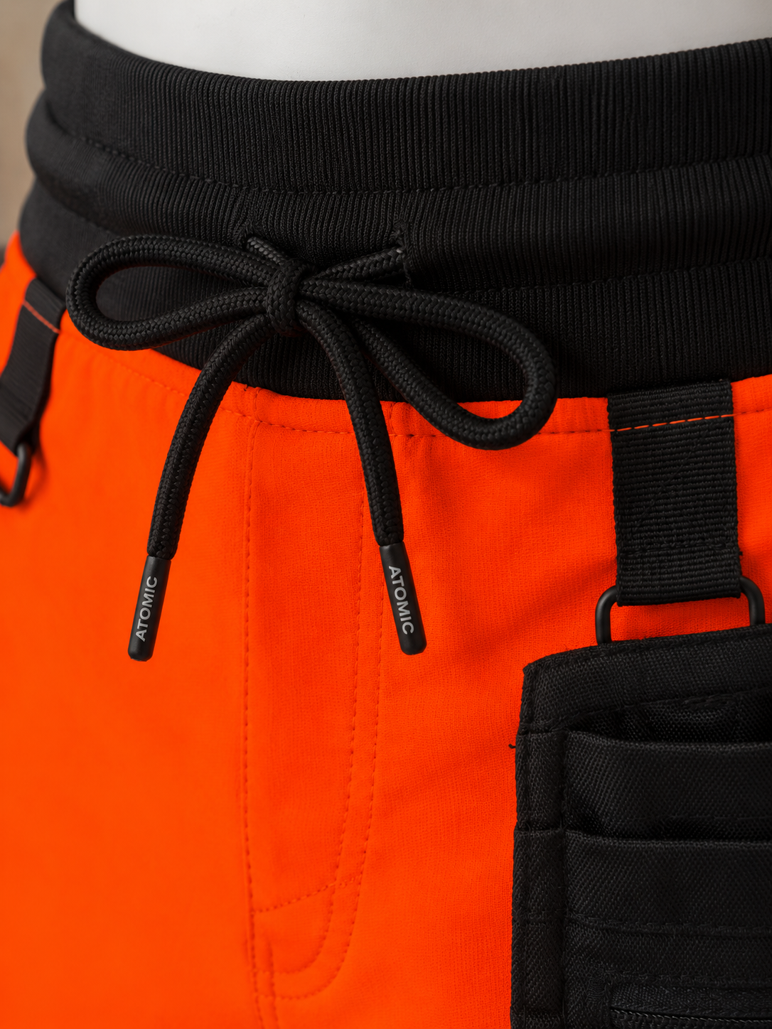 Close-up of bright orange athletic shorts with a black ribbed waistband, black drawstring, and black utility pocket on a mannequin.